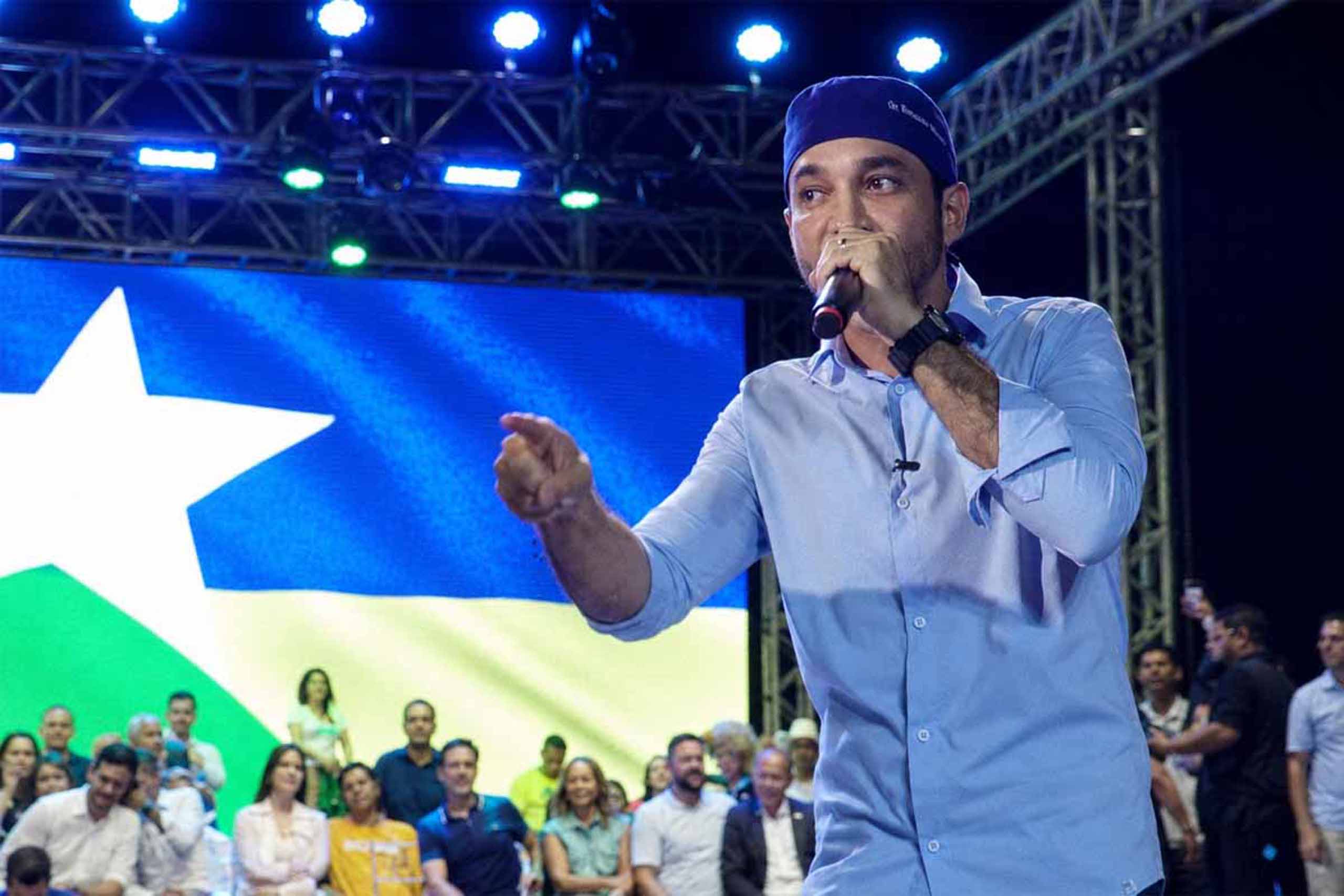 Homem de camisa azul fala ao microfone em palco diante de uma plateia